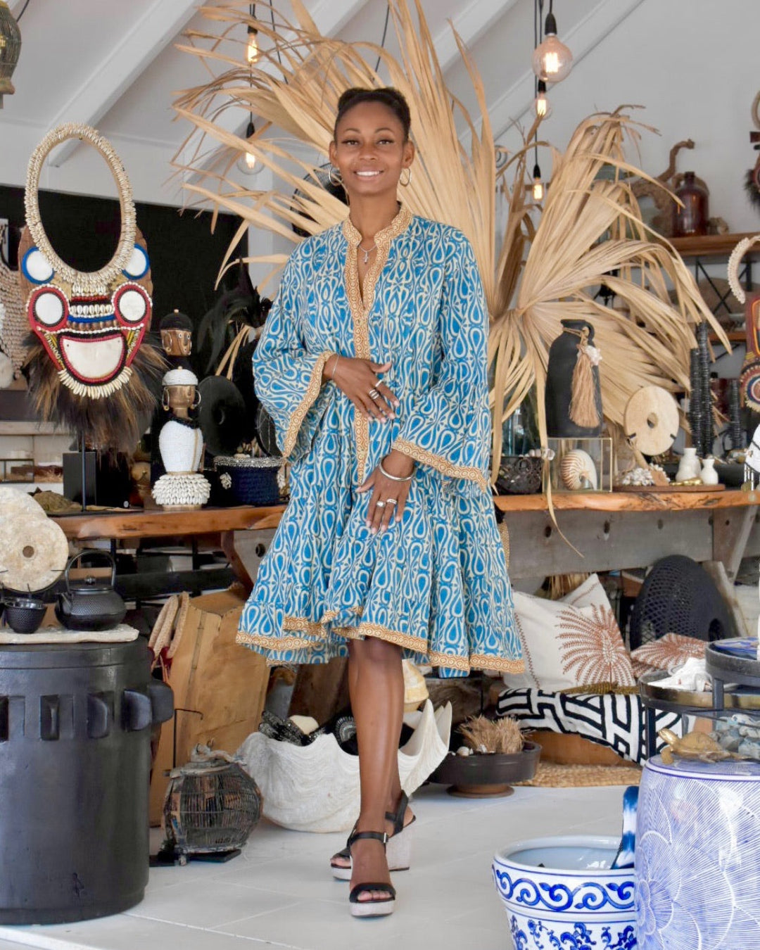 Woman in a blue patterned dress standing in a room with decorative items and plants.