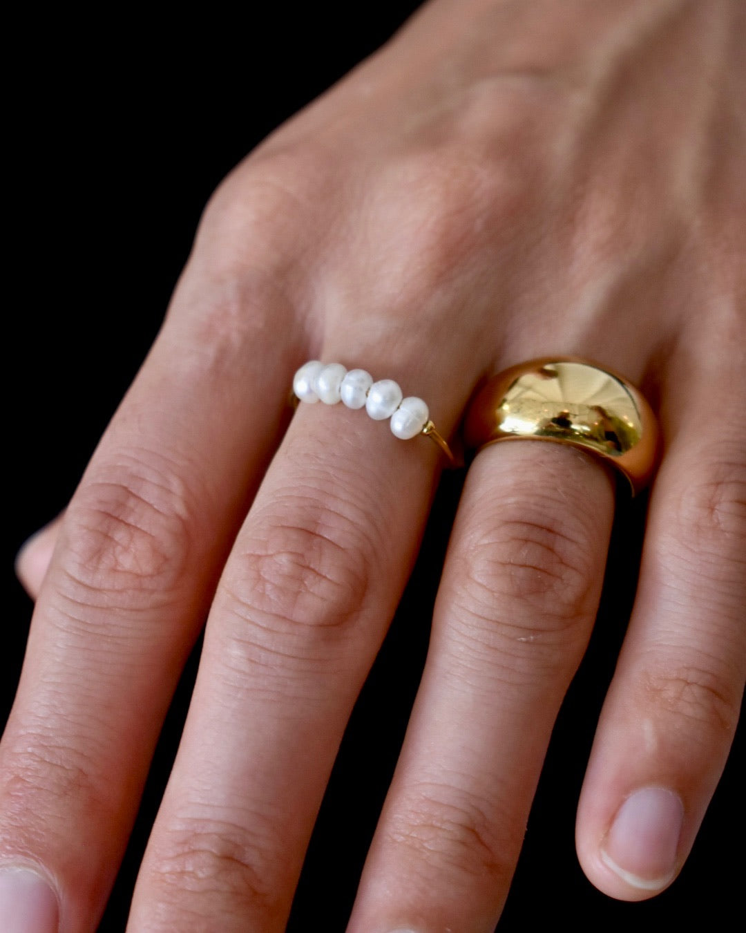 Hand wearing a gold ring and a pearl-studded ring on a black background
