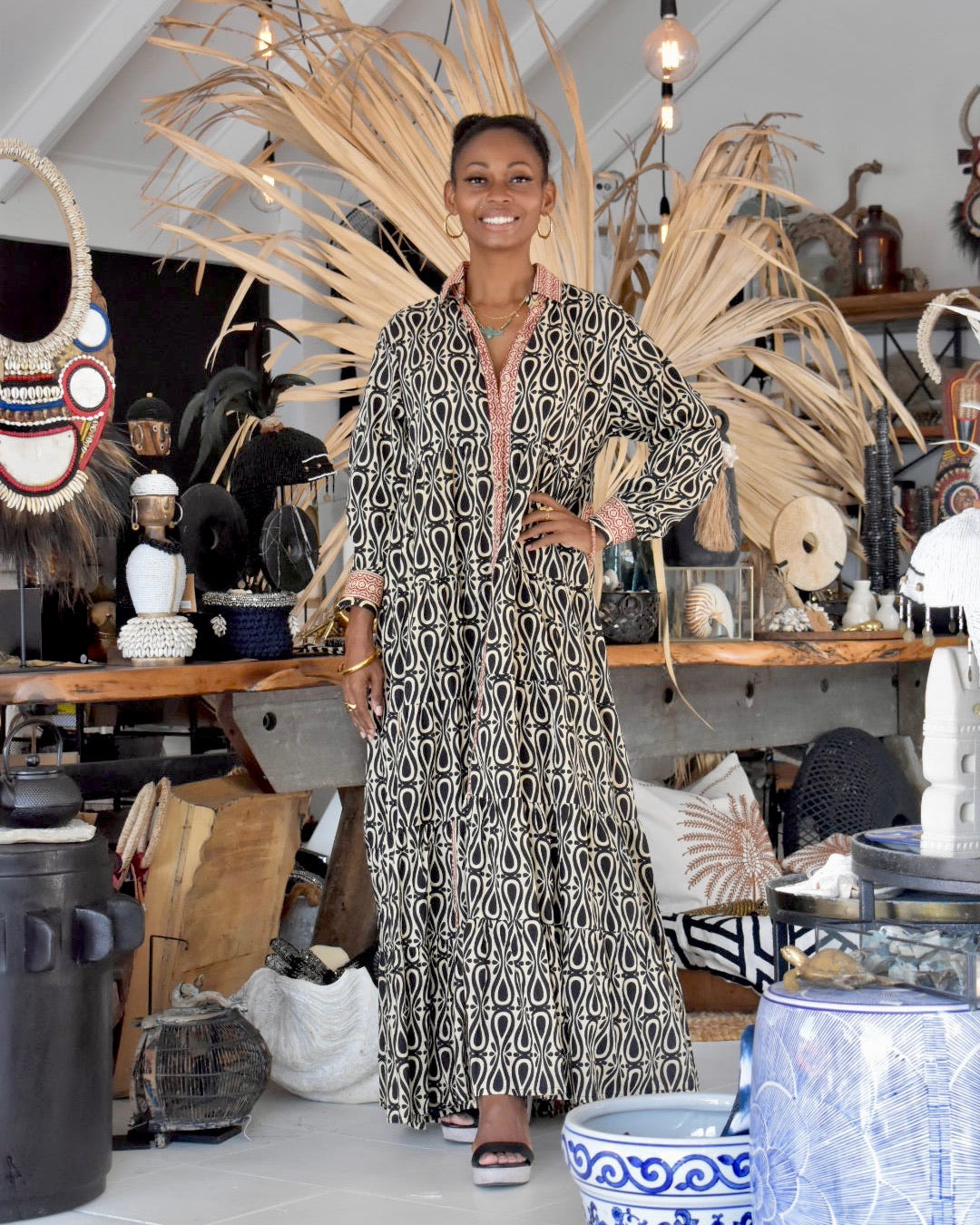 Woman in a patterned dress standing in an interior setting with decorative items.