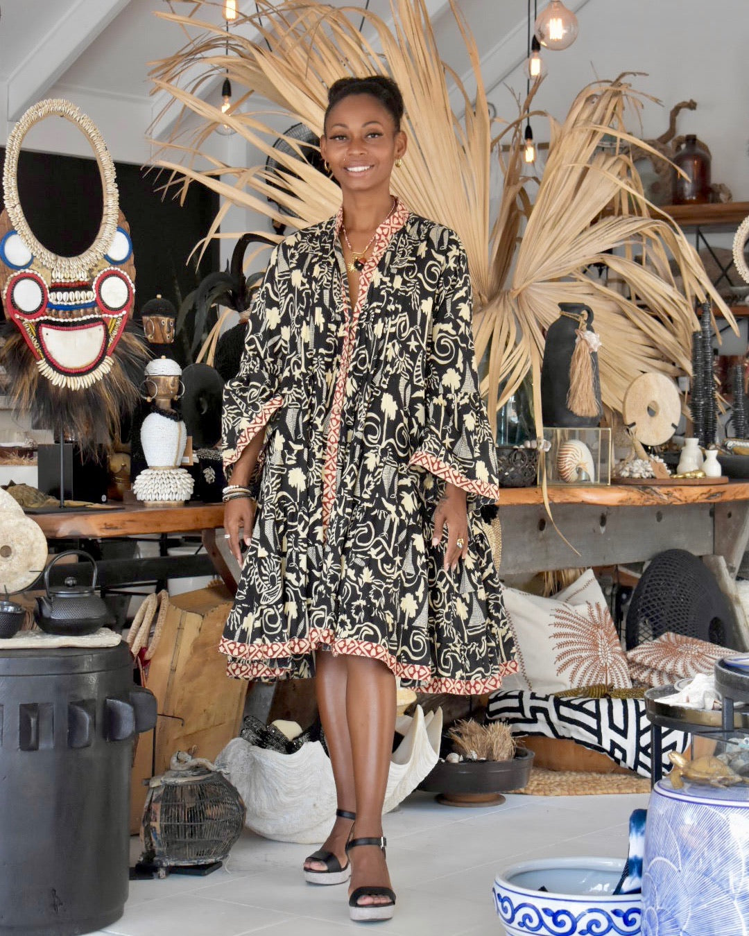 Woman in a patterned dress standing in a room with decorative items and plants.